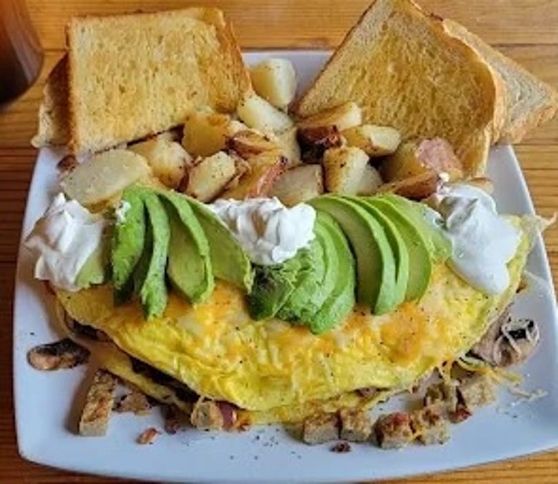 A fluffy omelet topped with avocado slices and sour cream, served with toast and roasted potatoes.