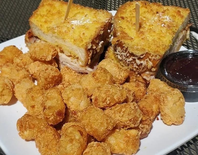 Two golden-brown Monte Cristo sandwiches with a side of tater tots and a dipping sauce.