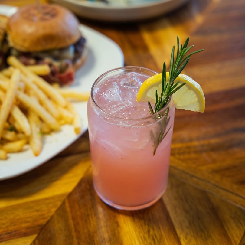 Pink cocktail with lemon and rosemary garnish, next to a plate of sliders and fries.