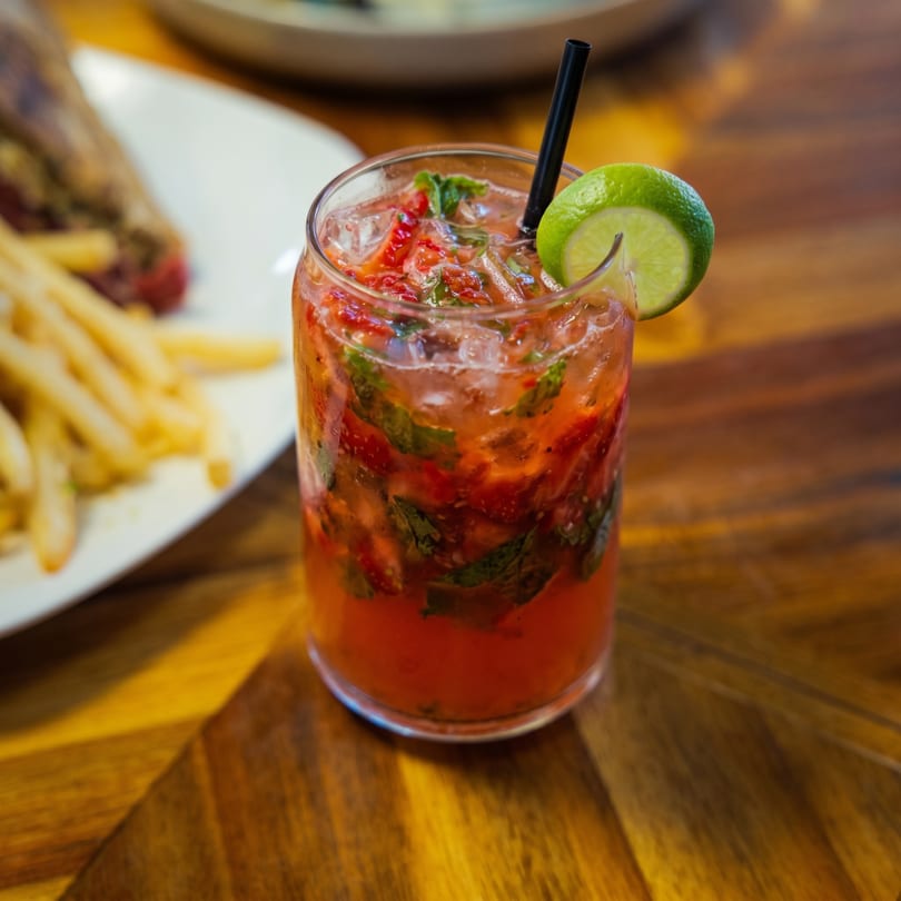 A strawberry mojito with a lime wedge and black straw sits on a wooden table next to fries.