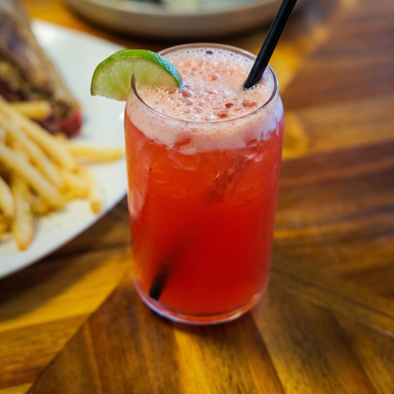 A vibrant red cocktail with foam and a lime wedge, next to a plate of fries.