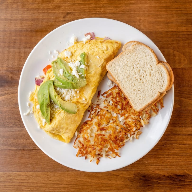 Omelet with avocado, feta, and red onion, served with hash browns and toast on a white plate.